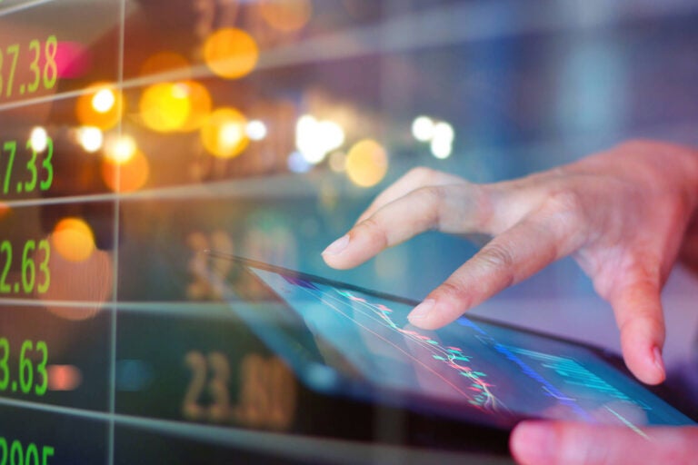 Image of hand working on tablet with financial charts and stock tickers in the background.